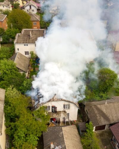 Aerial view of a house on fire with orange flames and white thick smoke.